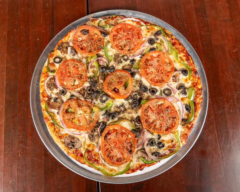 A whole vegetarian pizza with tomatoes, olives, mushrooms, onions, and green peppers on a metal pan.