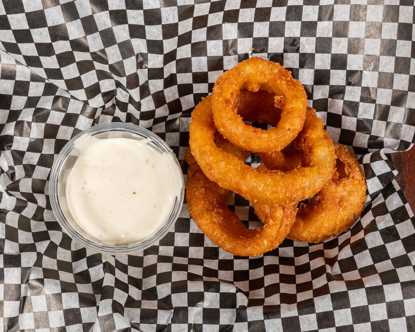 Golden-brown onion rings and a side of creamy dipping sauce on black and white checkered paper.