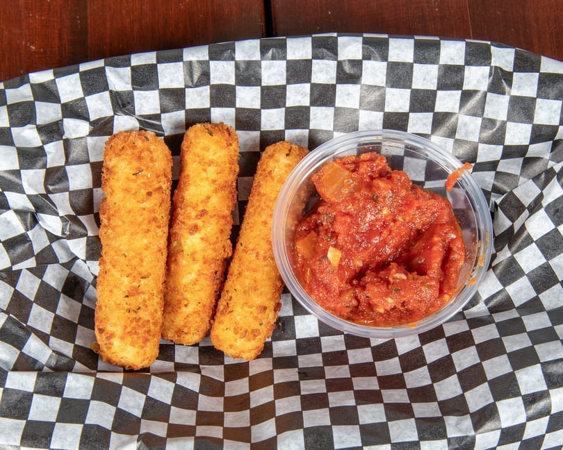 Three golden-brown mozzarella sticks and a clear cup of chunky marinara sauce in a checkered paper-lined basket.